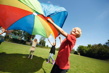 Kinder spielen mit einem großen, bunten Fallschirm auf einer Wiese unter einem klaren blauen Himmel. Ein Kind im Vordergrund hat ein rotes Hemd an, während andere Kinder im Hintergrund den Fallschirm halten. Das Feld ist von Bäumen gesäumt.