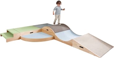 Ein kleines Kind steht auf einem hölzernen Indoor-Spielgerät mit Rampen, Stufen, Kurven und Plattformen. Die Oberfläche ist mit Teppich und Stoffteilen in Grün-, Blau- und Beigetönen bedeckt.