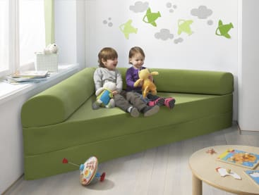 Zwei kleine Kinder sitzen auf einem grünen Sofa, jedes hält ein Stofftier. Auf dem hellen Holzboden sind Spielzeuggegenstände verstreut, und grüne Flugzeugaufkleber schmücken die weißen Wände im Hintergrund.