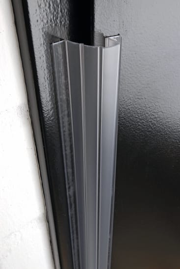 Nahaufnahme einer schwarzen Metalltür mit einer vertikalen grauen Kunststoffdichtung an der Kante, neben einer weißen Wand.