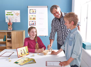Ein Mann und zwei Kinder beschäftigen sich an einem Tisch in einem Klassenzimmer mit Unterrichtsmaterial. Die Kinder lächeln. Im Hintergrund sind Karten, Modelle und ein Flipchart mit Diagrammen zu sehen.