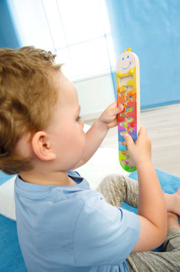 Ein kleines Kind mit hellbraunem Haar und blauem Hemd sitzt auf einem Bett und untersucht in einem hellen Zimmer ein buntes vertikales Spielzeug mit Schiebeperlen.