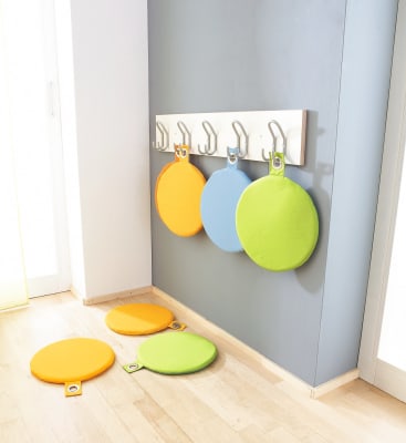 Vier runde, farbenfrohe Sitzkissen - gelb, orange, blau und grün - hängen an Metallhaken an einem Wandregal. Zwei gelbe und ein grünes Kissen liegen auf dem hellen Holzboden darunter.