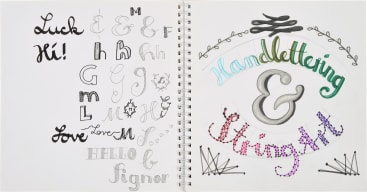 Ein aufgeschlagenes Skizzenbuch zeigt auf zwei Seiten verschiedene handgeschriebene Stile von Buchstaben, Wörtern und Symbolen, wobei der Schriftzug Handlettering & Stringart auf der rechten Seite in Farbe hervorgehoben ist.