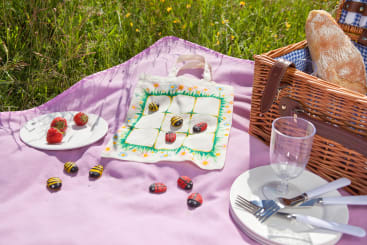 Ein Picknick auf einer Lavendeldecke mit einem Weidenkorb, Brot, Tellern, Besteck, Gläsern, Erdbeeren auf einem Teller und einer Serviette, die als Tic-Tac-Toe-Brett mit aufgemalten Steinbienen und Marienkäfern dient.