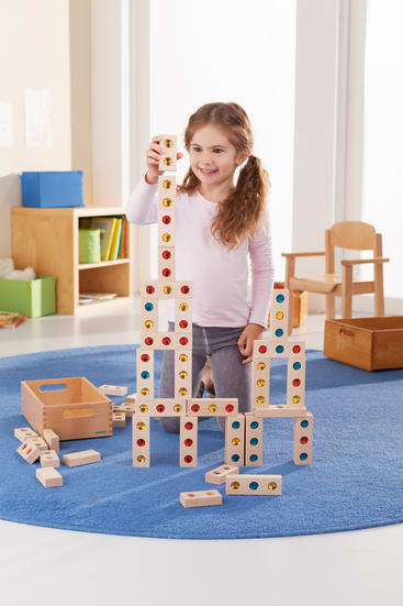 Ein junges Mädchen in einem langärmeligen Hemd und Jeans sitzt auf einem blauen Teppich und lächelt, während sie mit Holzklötzen, die farbige Kreise haben, ein hohes Bauwerk errichtet. Im Hintergrund sind Holzmöbel und Kisten zu sehen.
