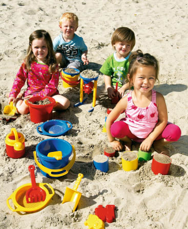 Vier kleine Kinder spielen im Sand mit bunten Plastikeimern, Schaufeln und anderem Strandspielzeug. Sie sitzen und knien im Sand, umgeben von verschiedenen Sandspielzeugen.