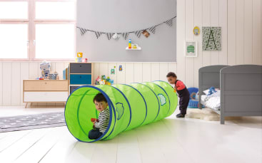 Zwei kleine Kinder spielen in einem Schlafzimmer. Ein Kind sitzt in einem grünen Spieltunnel und hält eine rote Tasse, während das andere Kind am Eingang des Tunnels steht. Das Zimmer ist mit Spielzeug, einem Bett und dekorativen Gegenständen an den Wänden ausgestattet.