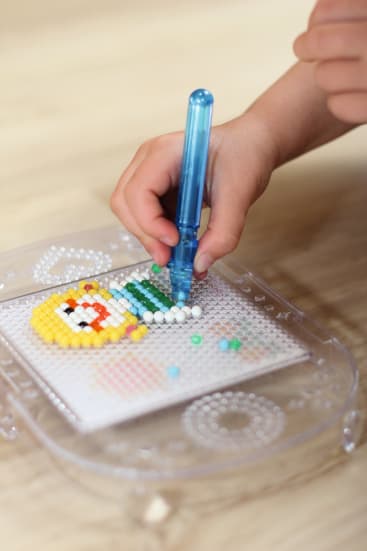 Eine Kinderhand setzt mit einem blauen Werkzeug kleine bunte Perlen auf ein Steckbrett, wodurch ein Perlenmuster auf einem transparenten Tablett entsteht, das auf einer hellen Holzfläche steht.
