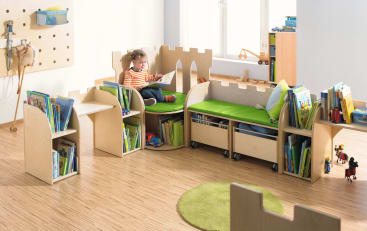 Ein kleines Kind sitzt lesend auf einer grün gepolsterten, schlossförmigen Holzbank, umgeben von Regalen mit Kinderbüchern in einem hellen, organisierten Spielzimmer mit Holzfußboden.