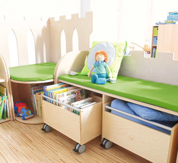 Eine Leseecke für Kinder mit grün gepolsterten Bänken, Holzschränken mit Büchern und Spielzeug, einem blauen Kissen und einer Plüschpuppe, die auf einem Kissen sitzt, alles auf einem hellen Holzboden.