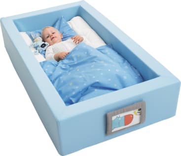 Ein Baby liegt in einem gepolsterten, rechteckigen blauen Kinderbett mit blauem Bettzeug und einem Kissen. Das Bettchen hat weiche Seiten und einen Stoffaufnäher mit einer Tierillustration auf der Vorderseite.