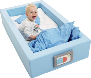 Ein lächelndes Baby sitzt aufrecht in einem Bett mit hellblau gepolsterten Seiten, das mit einem blauen Kissen und einer Decke mit kleinen weißen Mustern bedeckt ist. Eine kleine Tierillustration ziert das Ende des Bettes.