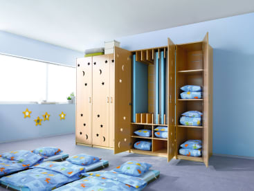 Ein Zimmer mit hellblauen Wänden, mehreren Schlafmatten mit Kissen und Decken auf dem Boden und einem großen Holzschrank, in dem gefaltete Bettwäsche und Schlafmatten aufbewahrt werden. Die Schranktüren sind mit Stern- und Mondausschnitten versehen.
