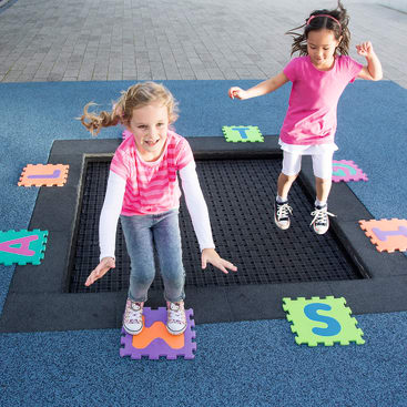 Zwei junge Mädchen springen auf einem in den Boden eingelassenen Trampolin, umgeben von bunten Buchstabenfliesen auf einem blauen Gummibelag in einem Spielbereich im Freien.