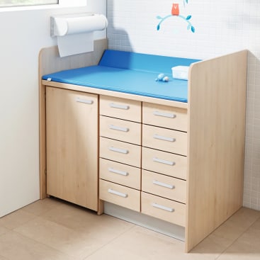 Ein hölzerner Wickeltisch mit mehreren Schubladen und einer Schranktür, darauf eine blaue gepolsterte Wickelauflage, ein kleiner weißer Behälter und eine Rolle Papierhandtücher. Eine weiß geflieste Wand und ein tierisches Wandtattoo im Hintergrund.
