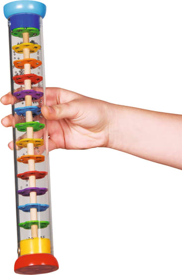 Eine Hand hält ein durchsichtiges zylindrisches Spielzeug mit einem Holzstab in der Mitte, mehreren bunten Plastikscheiben und kleinen Perlen im Inneren, das an einen Regenstab oder ein sensorisches Rohr erinnert.