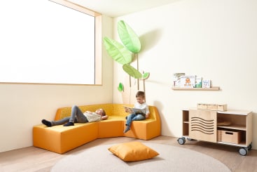 Zwei Kinder befinden sich in einem hellen Zimmer mit gelben Möbeln. Das eine liegt auf einem gelben Sofa, das andere sitzt und liest. Es gibt eine pflanzenförmige Wanddekoration, ein Fenster, ein kleines Bücherregal und einen runden Teppich.