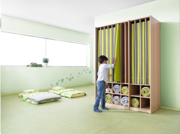 Ein kleines Kind in einem hellgrünen Zimmer steht an einem hölzernen Lagerschrank und zieht eine gefaltete grüne Matte heraus. Aufgerollte Decken und gestapelte Matten sind in dem Schrank aufgeräumt, und das Bettzeug liegt ordentlich auf dem Boden daneben.