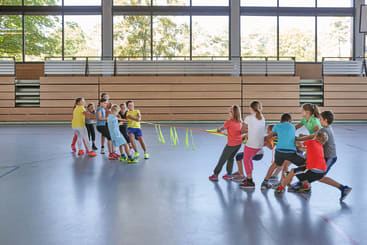 Eine Gruppe von Kindern spielt Tauziehen mit einem gelben Seil in einer Turnhalle. Die Turnhalle hat hölzerne Tribünen und große Fenster, aus denen man Bäume sehen kann.