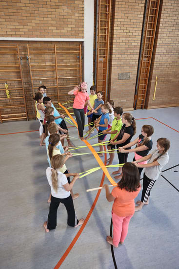 Eine Gruppe von Kindern und ein Erwachsener stehen in einer Turnhalle und halten jeweils bunte Bänder, die in der Mitte befestigt sind und einen Stern bilden. Der Erwachsene scheint die Aktivität zu leiten. Die Turnhalle hat Backsteinwände und Sportmarkierungen.