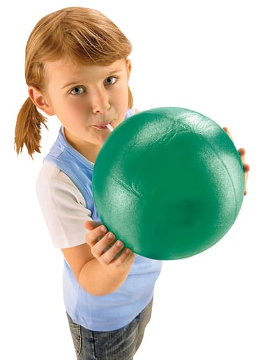 Ein junges Mädchen mit hellbraunem Haar in Zöpfen hält einen großen grünen Ball und blickt in die Kamera. Sie trägt ein blau-weißes Hemd und eine blaue Jeans und steht vor einem weißen Hintergrund.