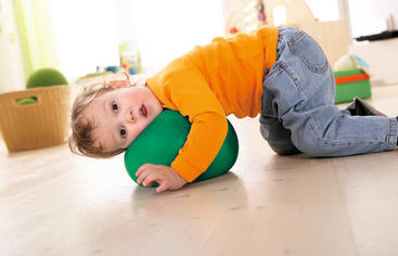 Ein kleines Kind in einem orangefarbenen langärmeligen Hemd und blauen Jeans liegt auf dem Boden und umarmt eine große grüne Schaumstoffform. Die Umgebung scheint ein heller, aufgeräumter Innenraum mit einem Korb und Spielzeug im Hintergrund zu sein.