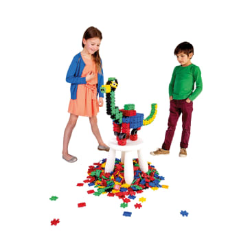 Zwei Kinder stehen neben einem weißen Hocker, auf dem ein bunter Dinosaurier aus Plastikbausteinen steht, und weitere Bausteine sind um den Hocker herum auf dem Boden verstreut.