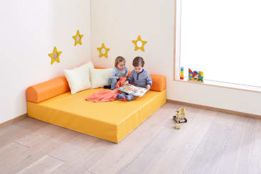 Zwei kleine Kinder sitzen auf einer orangefarbenen Matratze mit einer Decke und Kissen und lesen ein Buch. Das Zimmer hat einen Holzboden, gelbe Sterne an der Wand und Spielzeug in der Nähe eines großen Fensters.