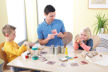 Ein Erwachsener und zwei Kinder sitzen an einem Tisch mit Unterrichtsmaterialien, darunter Bildkarten mit Gesichtszügen, Holzklötze und bunte Objekte. Der Erwachsene hält eine Karte mit Lippen, während die Kinder mit den Materialien interagieren.