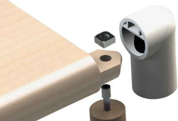 Eine hölzerne Tischplatte mit einer abgerundeten Kante hat ein Loch für eine Schraube. In der Nähe befinden sich ein Verbindungsblock aus Metall, eine teilweise im Holz befestigte Schraube und ein hohler, zylindrischer Eckbeschlag aus weißem Kunststoff.