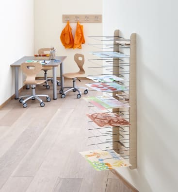 Ein Klassenzimmer mit zwei Tischen und Stühlen, orangefarbenen Kitteln, die an Haken hängen, Malutensilien auf den Tischen und ein an der Wand befestigter Trockenständer mit mehreren Kinderbildern und -zeichnungen.