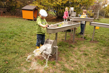 Drei Kinder spielen an einem Wassertisch im Freien auf einer Wiese. Ein Junge dreht ein Rad, so dass Wasser herausfließt, während ein Mädchen und ein anderer Junge im Hintergrund mit Wasserspielzeug spielen. Bäume und ein kleiner roter Schuppen sind zu sehen.