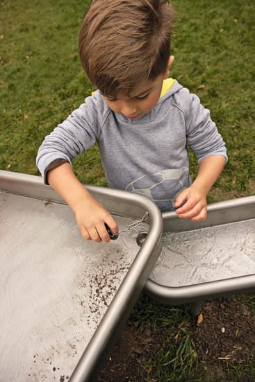 Ein kleines Kind in einem grauen Kapuzenpulli spielt mit Wasser auf einem metallenen Wassertisch im Freien, wobei es ein Drahtwerkzeug benutzt. Im Hintergrund ist grünes Gras zu sehen.