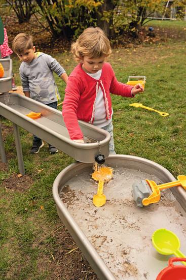 Ein kleines Kind in einem rosa Pullover spielt an einem Wassertisch im Freien mit Spielzeugschaufeln und Eimern. Ein anderes Kind steht in der Nähe. Die Szene spielt auf einer Wiese mit Bäumen und Spielzeug im Hintergrund.