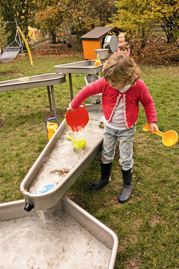 Ein kleines Kind in einer rosa Jacke und Regenstiefeln schüttet Wasser aus einem roten Behälter in einen Wasserspieltisch im Freien. Ein anderes Kind und Spielgeräte sind im Hintergrund zu sehen. Gras und Herbstblätter liegen auf dem Boden.