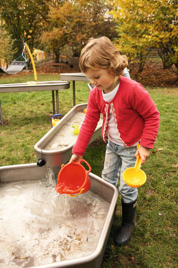 Ein kleines Kind in einer rosa Jacke und schwarzen Stiefeln gießt Wasser aus einem roten Eimer in ein Metallbecken an einer Wasserspielstation im Freien auf einer Wiese, mit Bäumen und Spielgeräten im Hintergrund.