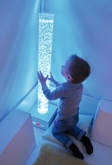Ein kleiner Junge kniet in einem blauen, sanft beleuchteten Raum auf Kissen, berührt eine hohe, zylindrische, mit Wasser gefüllte und von innen beleuchtete Seifenblasenröhre und schaut zu ihr auf.