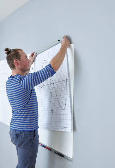 Eine Person in einem blau gestreiften Hemd hängt ein großes Blatt Millimeterpapier mit einem gezeichneten Diagramm an ein Whiteboard. Auf dem Tablett unter dem Whiteboard befinden sich einige Stifte und Marker.