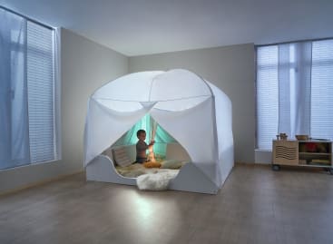 Ein Kind sitzt in einem weißen Innenzelt mit lichtdurchlässigen Vorhängen, umgeben von Kissen und einem flauschigen Teppich, in einem minimalistischen Raum mit großen Fenstern und Holzfußboden.