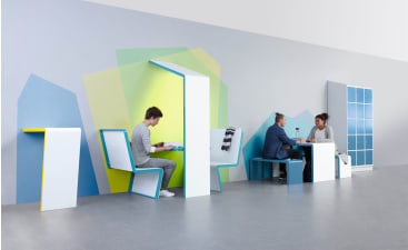 Drei Personen nutzen moderne, minimalistische Arbeitsplätze an einer Wand mit bunten geometrischen Mustern; eine Person arbeitet allein an einem Schreibtisch, während zwei Personen an einem Tisch mit Sitzbank ein Gespräch führen.
