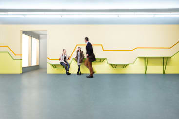 Ein Mann geht an zwei sitzenden Frauen in einem modernen, minimalistischen Raum mit gelben Wänden, geometrischen farbigen Linien und eingebauten Bänken vorbei. Der Raum hat einen blaugrauen Boden und ein beleuchtetes Fenster auf der linken Seite.