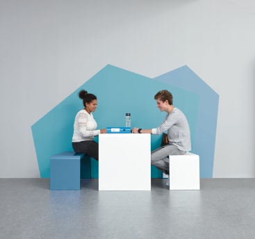 Zwei Personen sitzen sich an einem weißen Tisch gegenüber, jede auf einem blauen oder weißen Würfelsitz. Auf dem Tisch befinden sich eine Wasserflasche und ein Buch. Im Hintergrund sind geometrische blaue Formen auf einer grauen Wand zu sehen.