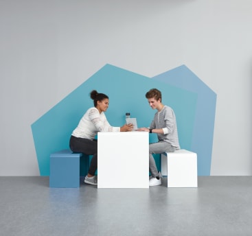 Zwei Personen sitzen sich an einem weißen Tisch auf Würfelsitzen gegenüber und halten eine Tasse. Im Hintergrund sind geometrische blaue Formen auf einer hellgrauen Wand zu sehen. Der Boden ist grau, und die beiden Personen scheinen in ein Gespräch vertieft zu sein.