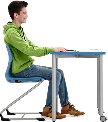 Eine junge Person in grünem Kapuzenpulli und Jeans sitzt aufrecht an einem blauen Schreibtisch, den Blick nach vorne gerichtet und die Hände auf den Tisch gestützt. Vor ihr liegt ein aufgeschlagenes Notizbuch. Der Hintergrund ist einfarbig weiß.