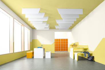 Ein heller, moderner Raum mit gelben und weißen geometrischen Wänden, grauem Fußboden und rechteckigen Deckenplatten. Er ist minimalistisch eingerichtet, mit Bänken, einem Tisch, Hockern und orangefarbenen Schränken im hinteren Bereich.