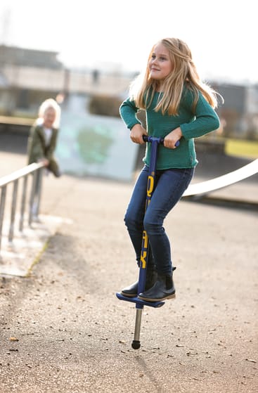 Ein junges Mädchen in grünem Pullover und Jeans benutzt einen blauen Pogo-Stick im Freien auf einer gepflasterten Fläche. Ein anderes Kind steht im Hintergrund in der Nähe eines Metallgeländers.