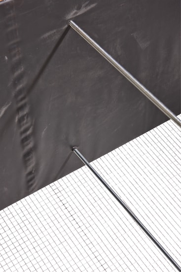 Ein schwarzes vertikales Paneel wird von zwei abgewinkelten Metallstäben getragen und liegt auf einer weißen, gitterartig gemusterten Fläche. Das Bild ist von oben aufgenommen und zeigt die geometrische Anordnung der Elemente.