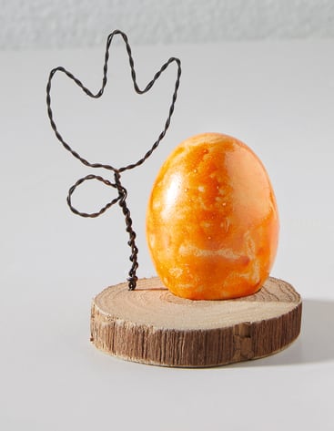 Eine orangefarbene Kugel sitzt auf einem runden Holzsockel neben einem Draht in Form eines Kaktus. Die Objekte sind auf einer weißen Fläche mit hellgrauem Hintergrund angeordnet.