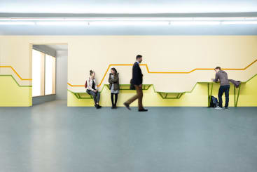 Drei Personen sitzen auf minimalistischen Bänken, die mit geometrischen, farbigen Linien an einer Wand befestigt sind, während eine weitere Person an einem hohen Tisch mit einem Rucksack auf dem Boden steht; zwei weitere Personen gehen unscharf in Bewegung vorbei.